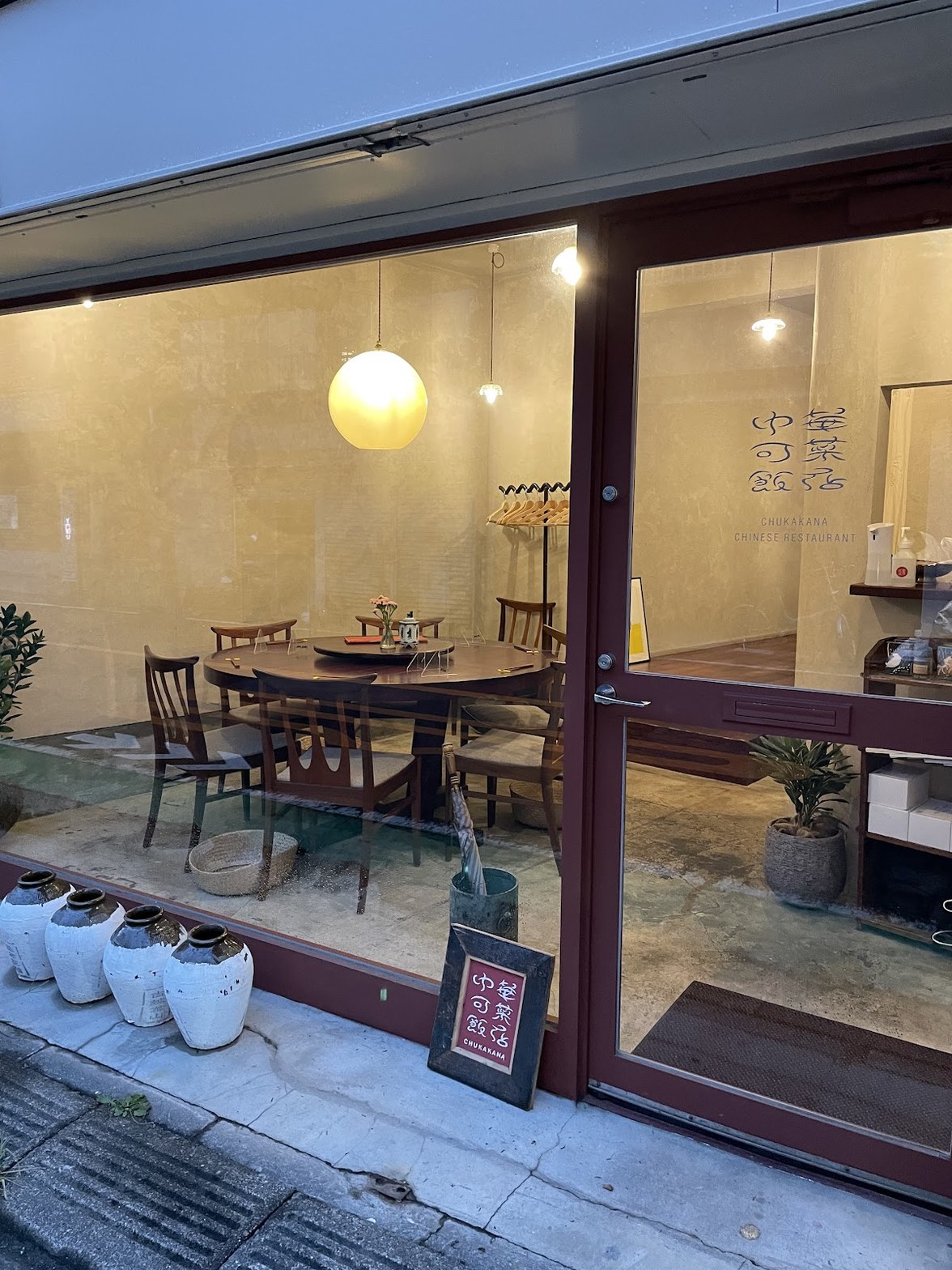 ガラス越しに見える店内の7人掛け円卓と丸い球形ペンダントライト、壁に筆文字で書かれた「中華可菜飯店」のロゴ